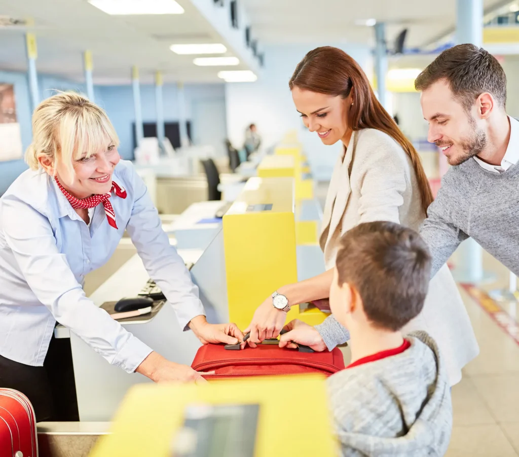 A smiling airline agent assists a family with a red suitcase at an airport check-in counter.